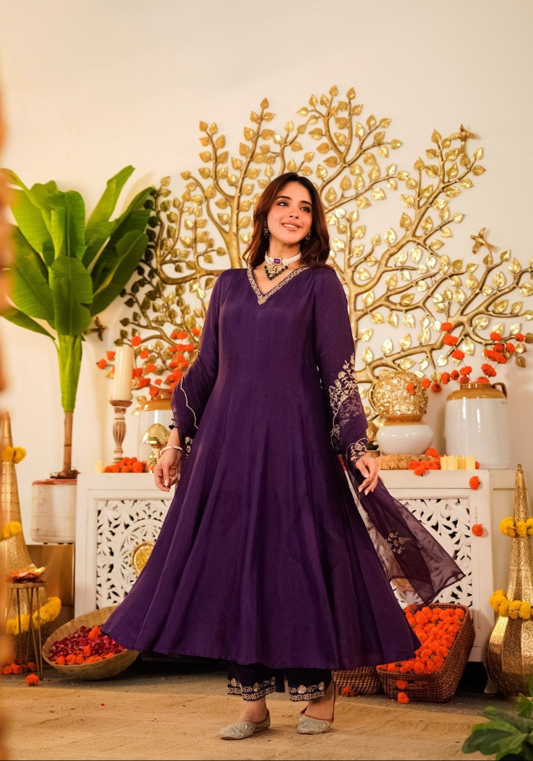 Woman in a purple dress standing in a decorated room with plants and decorative items.