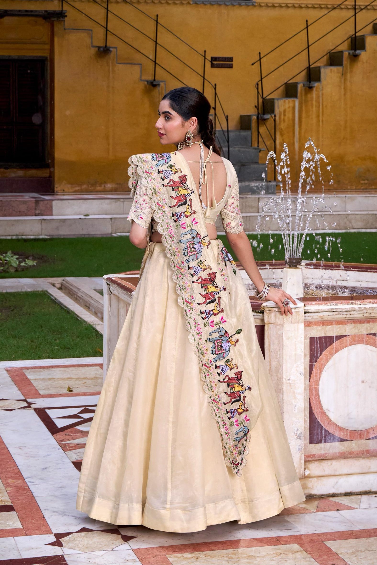 Woman in a traditional outfit with an embroidered sash standing in front of a fountain.