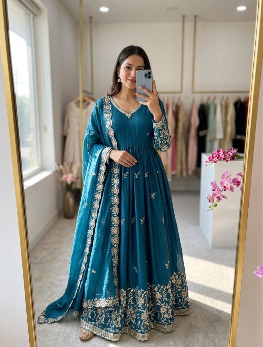 Woman taking a mirror selfie wearing a blue embroidered traditional outfit in a room with floral decorations.