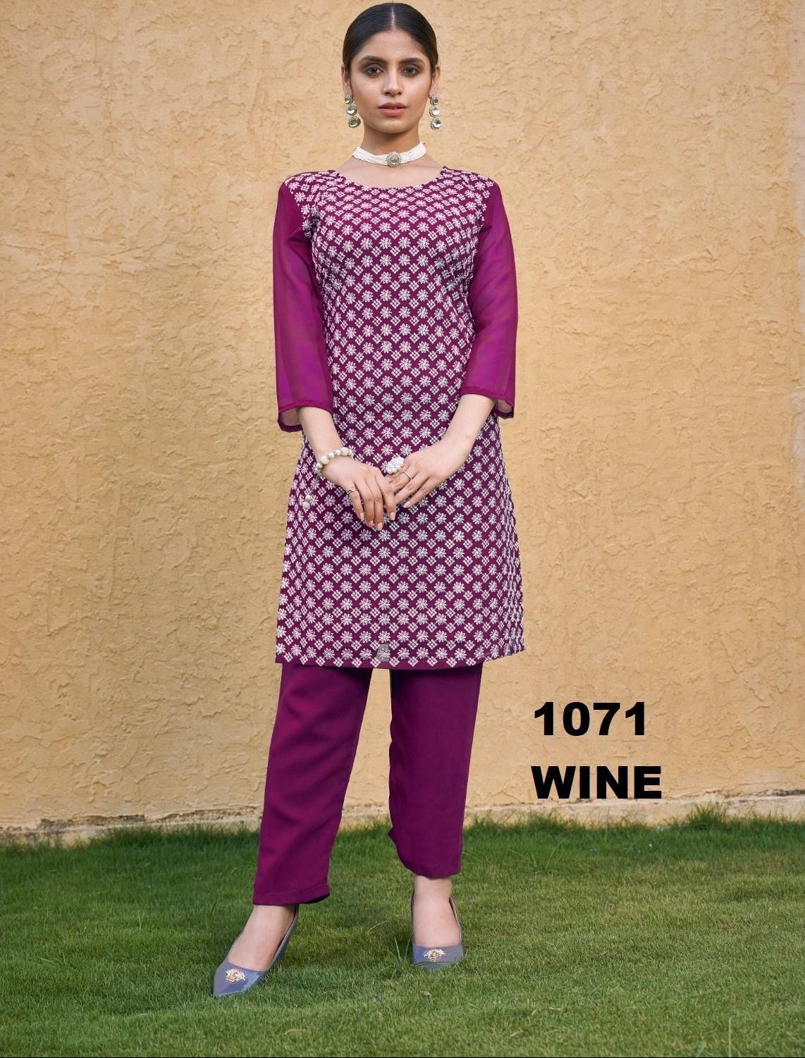 Woman wearing a purple outfit with a patterned top and pants on a grassy area with a beige wall background.