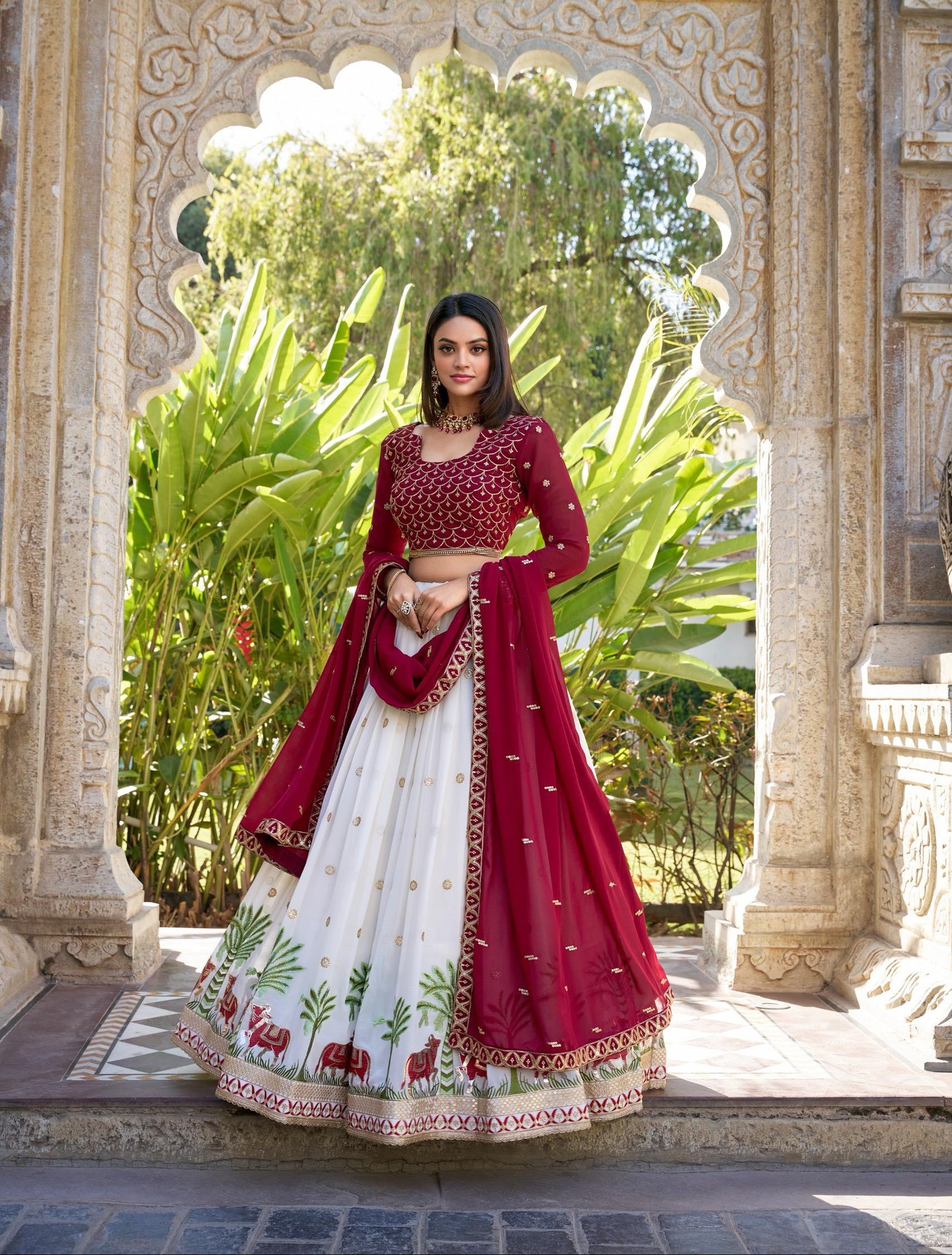 Woman in a red and white traditional outfit standing in front of an ornate archway with greenery.