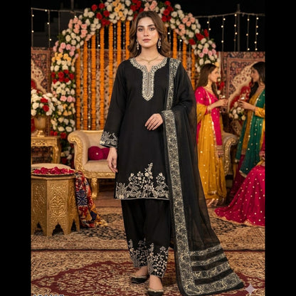 Woman in black embroidered traditional outfit standing in a decorated indoor setting with floral arrangements and people in colorful attire.