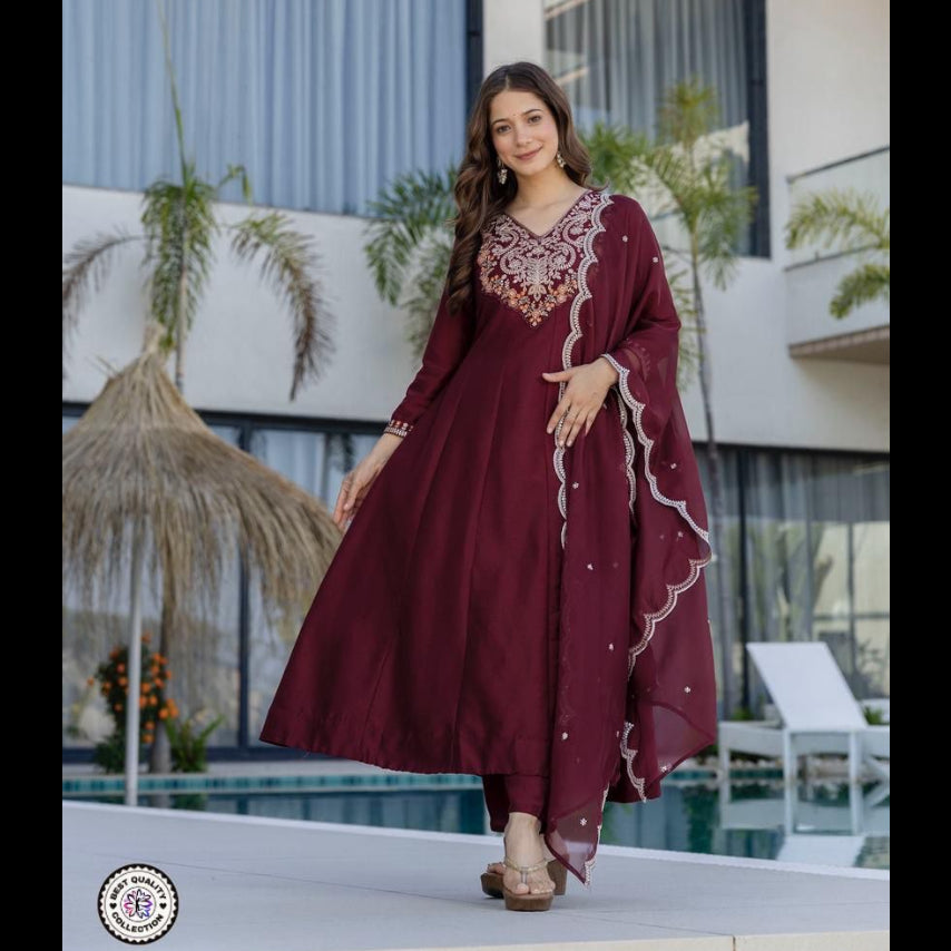 Woman in a burgundy dress with intricate designs standing in front of a modern building with plants.