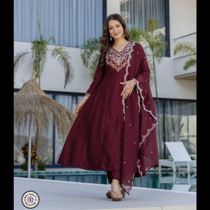 Woman in a burgundy dress with intricate designs standing in front of a modern building with plants.