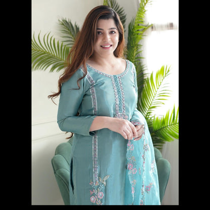 Woman wearing a light blue embroidered outfit sitting in a chair with green plants in the background