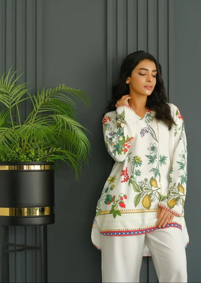 Woman wearing a floral blouse and white pants standing against a dark wall with a plant. (MARTIVA)