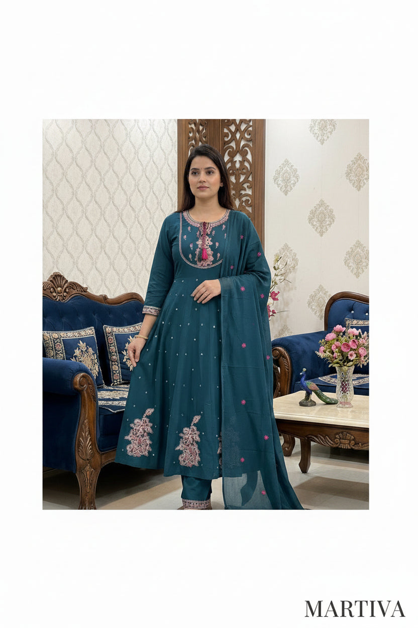 Woman in a teal embroidered outfit standing in a decorated room with furniture and decor.