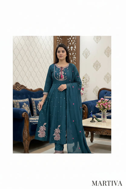 Woman in a teal embroidered outfit standing in a decorated room with furniture and decor.