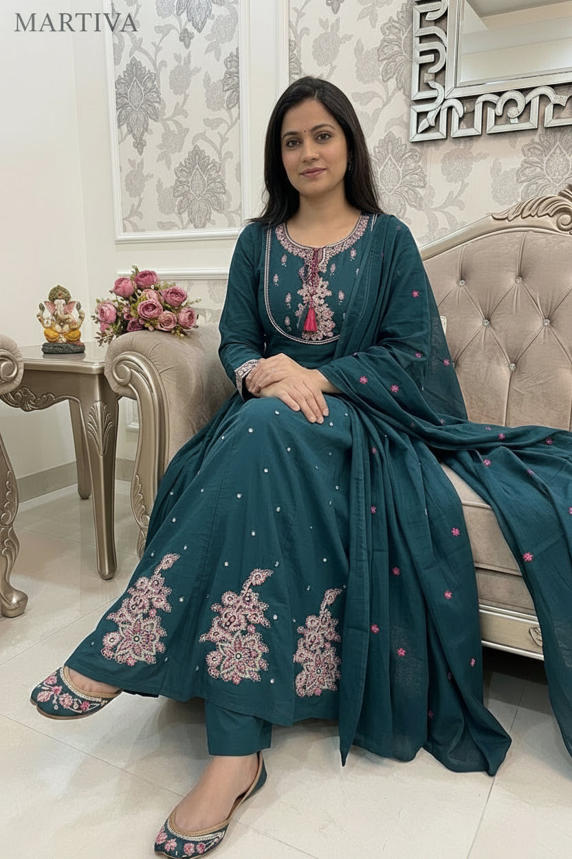 Woman in teal embroidered dress sitting on a sofa in a decorated room.