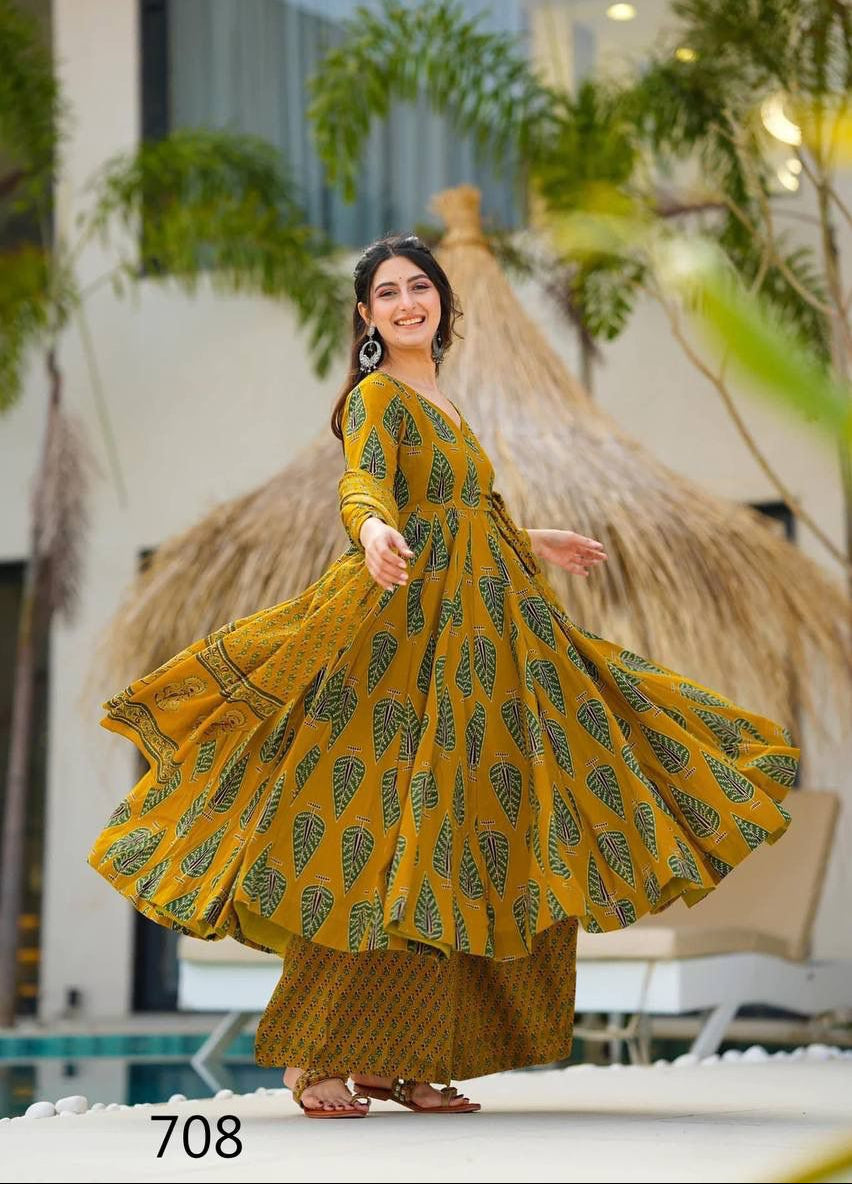 Woman in a yellow and green patterned dress standing outdoors with plants and a building in the background.