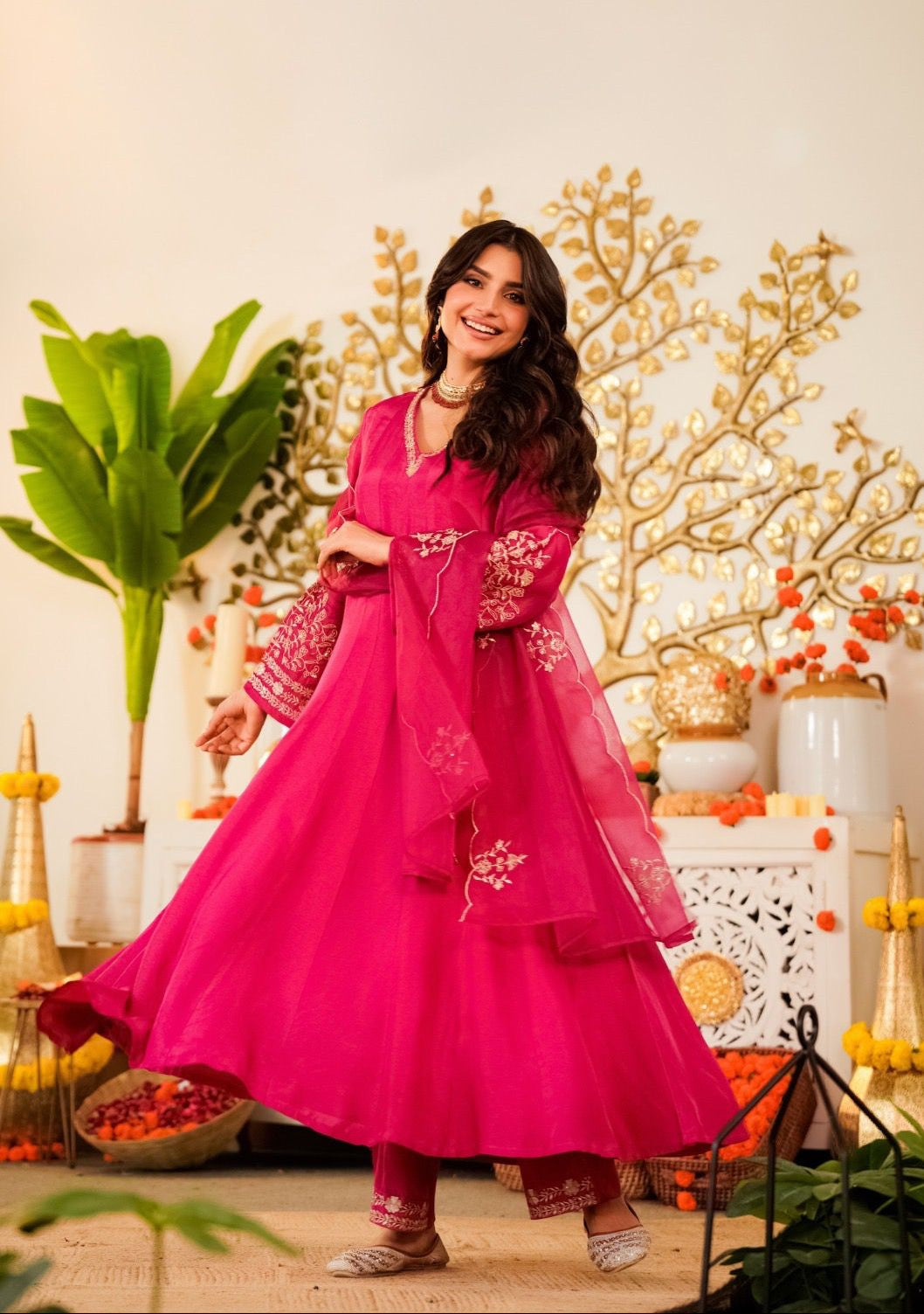 Woman in a pink traditional outfit standing in a decorated room with plants and decorative items.