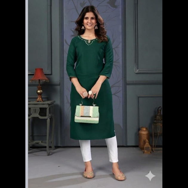 Woman wearing a green dress with white pants, holding a handbag, against a gray wall.