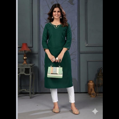 Woman wearing a green dress with white pants, holding a handbag, against a gray wall.