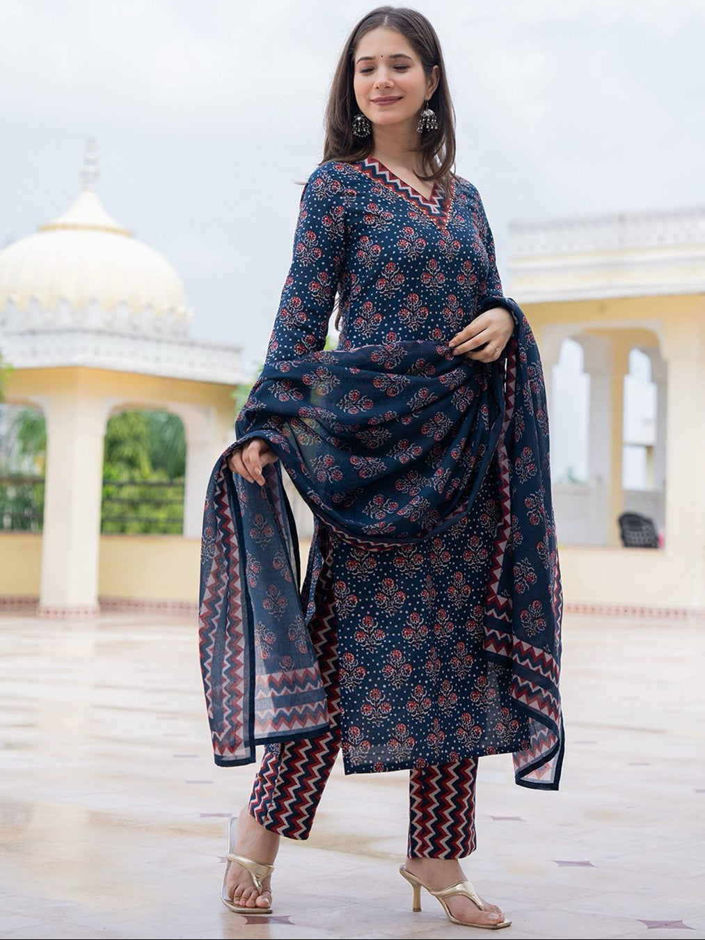 Woman in a traditional outfit with a patterned dupatta standing in front of a white architectural background.