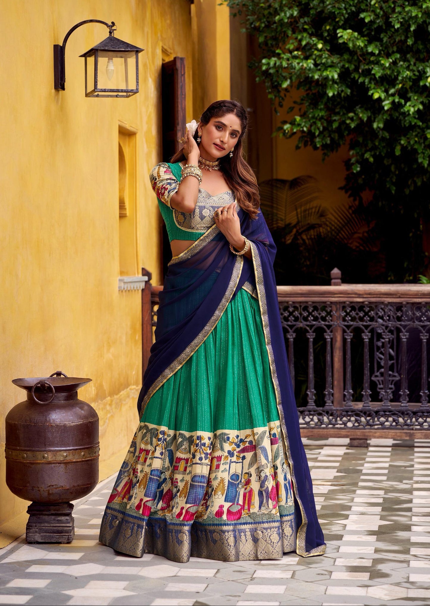 Woman in a traditional green and blue outfit standing in front of a yellow wall with a decorative lantern.