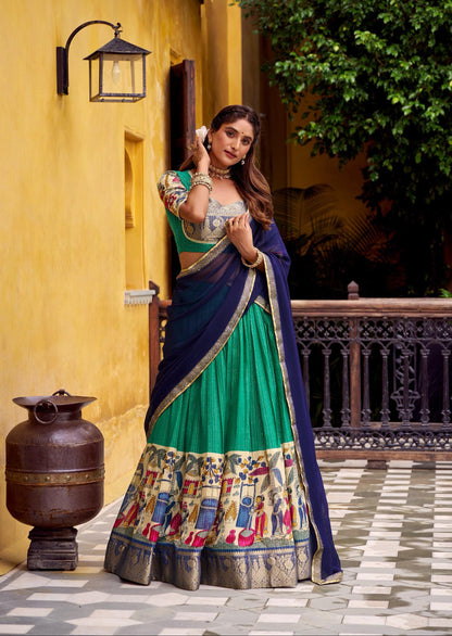 Woman in a traditional green and blue outfit standing in front of a yellow wall with a decorative lantern.