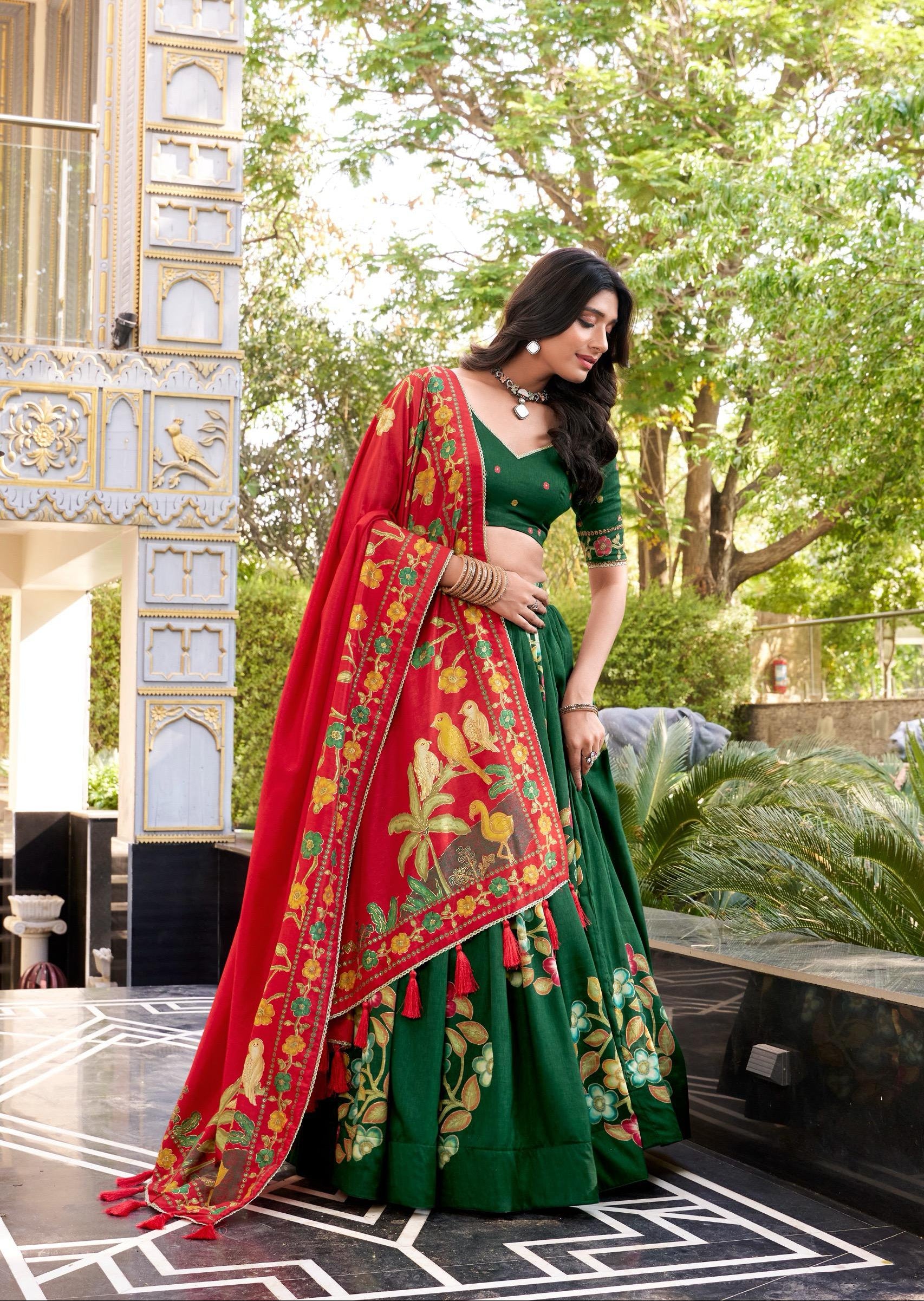Woman in a green and red traditional outfit standing outdoors with trees and architectural elements in the background.
