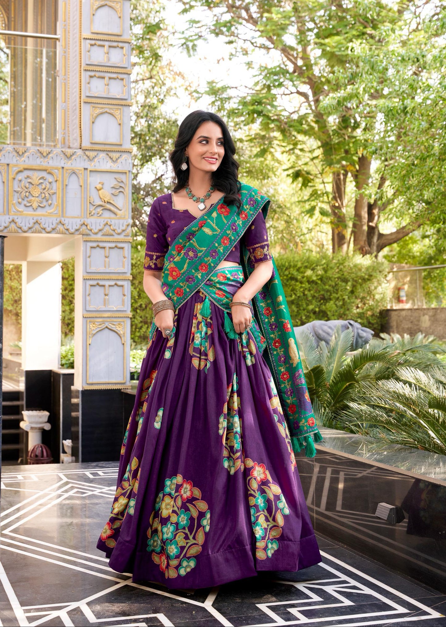 Woman in a purple traditional outfit with a green and red saree, standing outdoors.
