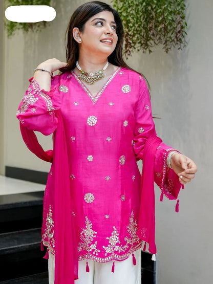 Woman wearing a bright pink embroidered traditional outfit with white pants, standing outdoors.